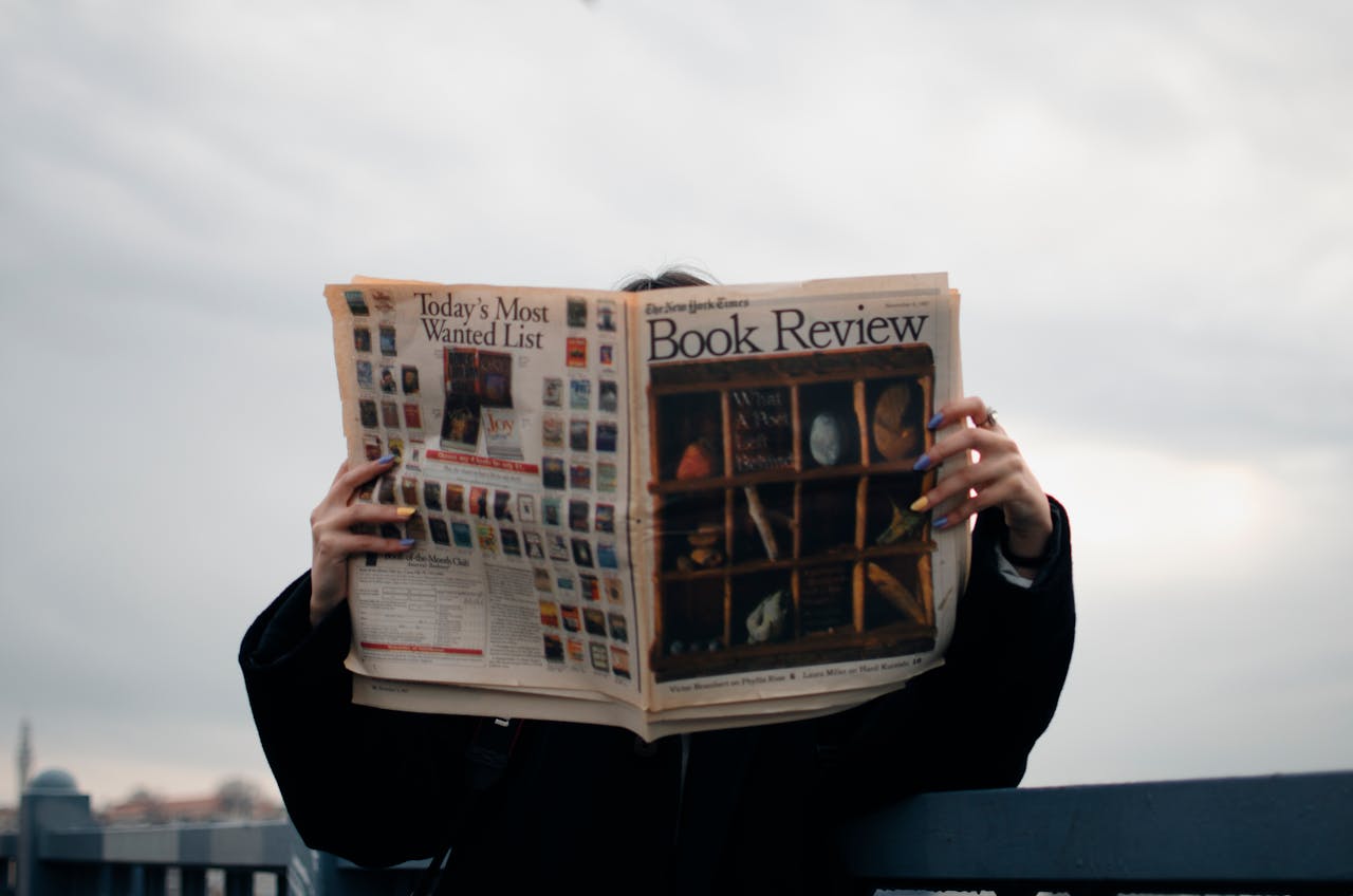 services-04 Individual holding a book review newspaper with colorful nails against a cloudy sky backdrop.