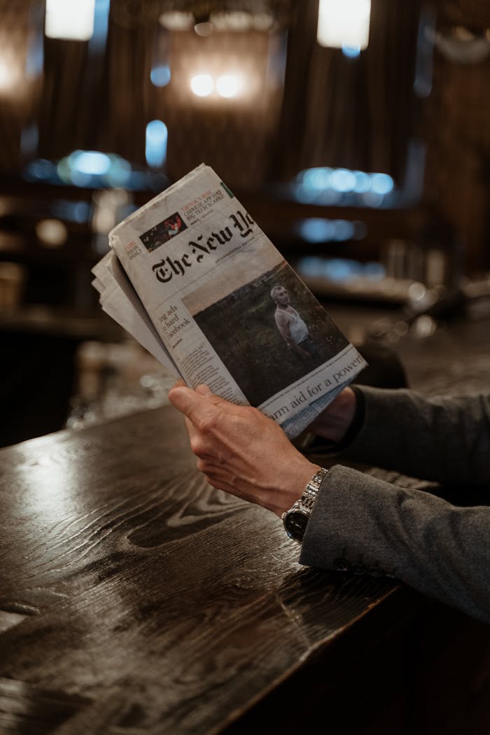 who-we-are Man holding a newspaper at a wooden bar table in a cozy, dimly lit setting.